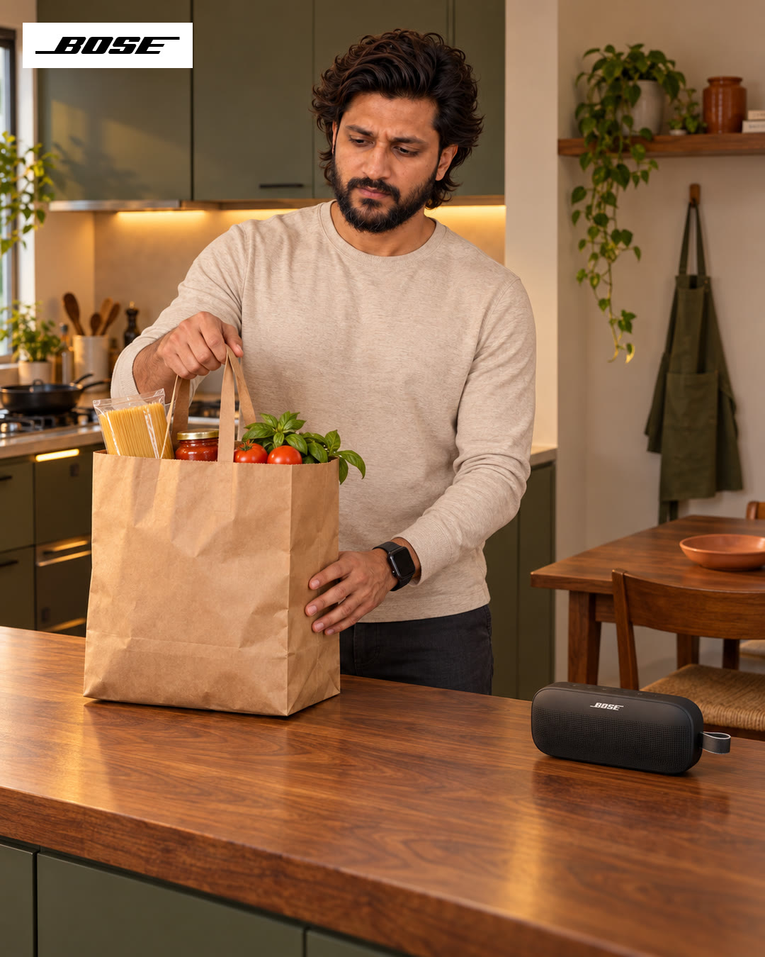 Man in a cream sweater unpacking a paper grocery bag on a wooden kitchen counter, a Bose speaker beside him.
