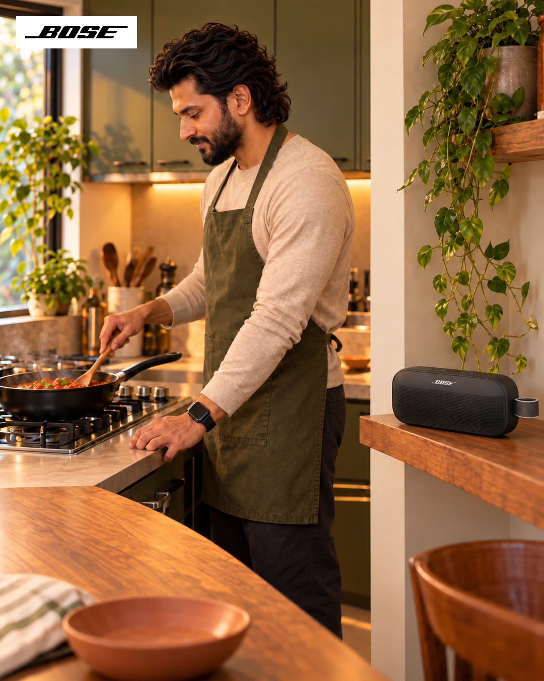 Man in an olive apron stirring a pan on the stove, the Bose speaker on the counter.
