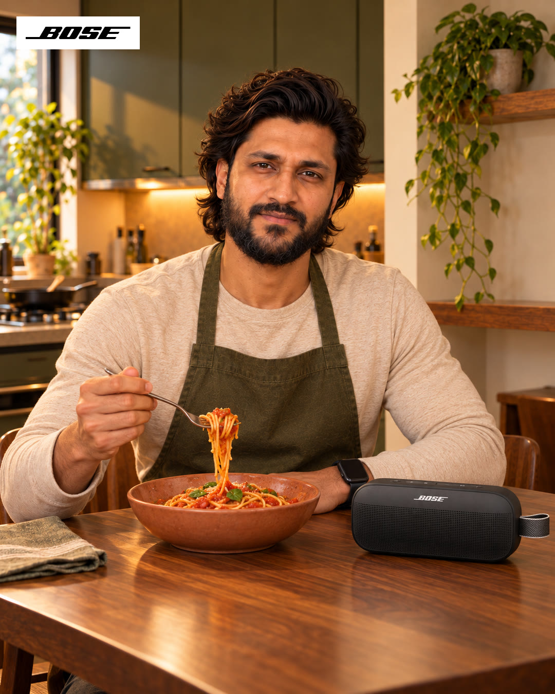 Man in an olive apron sitting at the table with a fork of pasta, a Bose speaker beside the bowl.