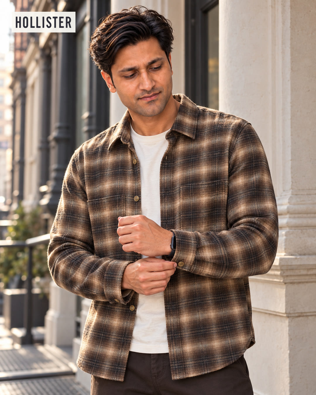 Man in a brown plaid flannel shirt over a white tee, looking down as he buttons his cuff.