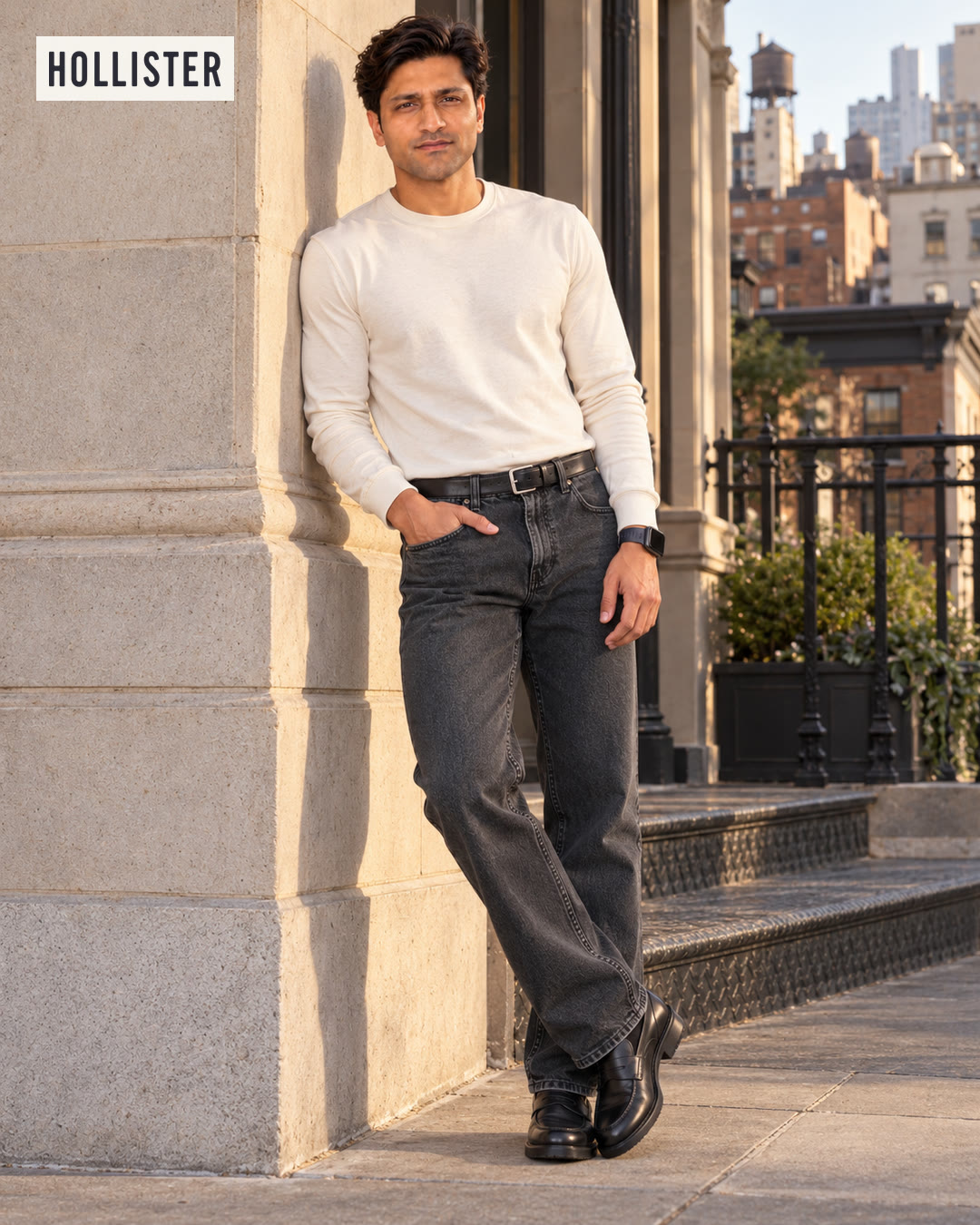 Man in a cream long-sleeve tee and washed black jeans leaning against a stone column with city rooftops behind him.