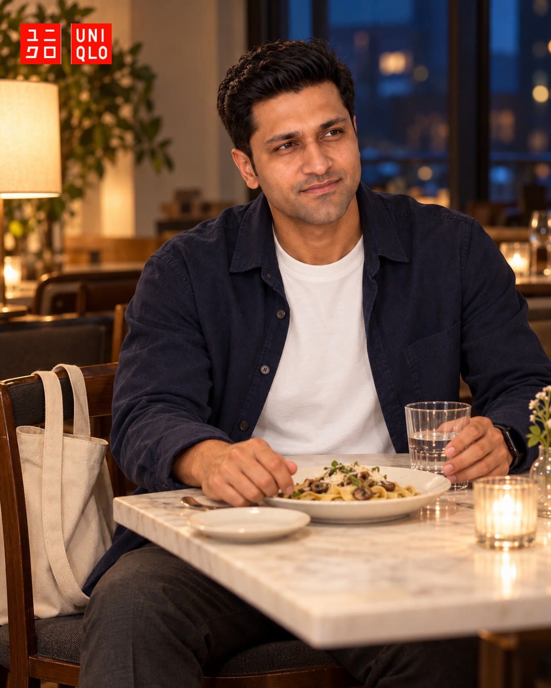 Man in a navy overshirt over a white t-shirt at a candlelit restaurant table with a plate of pasta.